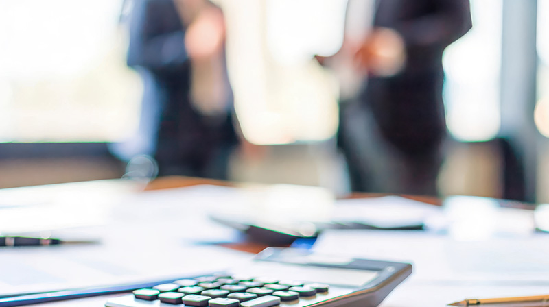 Table of documents with a calculator and pens and two people speaking in the background